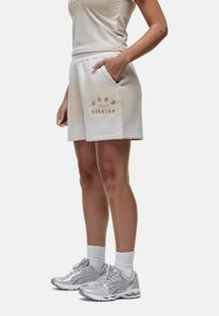 Pantaloni corti in cotone beige chiaro con vestibilità rilassata, tasche laterali e testo decorativo. Abbinati a scarpe sportive argentate e calze bianche lunghe.