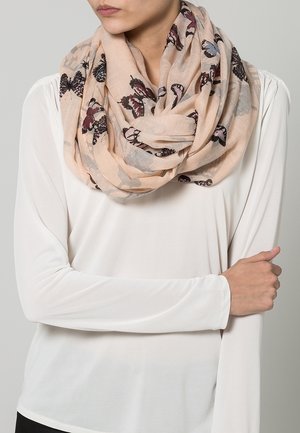 Vrouw draagt een licht beige oneindige sjaal met vlinderprint over een wit shirt met lange mouwen, armen gekruist ter hoogte van de taille tegen een neutrale achtergrond.