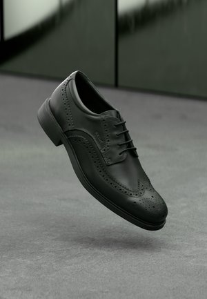 Czarny skórzany but brogue z detalami typu wingtip i sznurowadłami, zawieszony nad szarą podłogą i tle ciemnozielonej ściany.