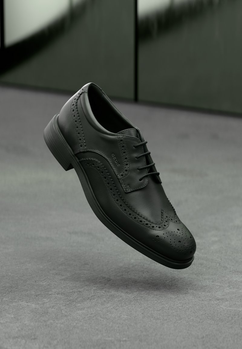 Chaussure brogue en cuir noir avec détails en bout d'aile et lacets, suspendue au-dessus d'un sol gris et d'un mur vert foncé en arrière-plan.
