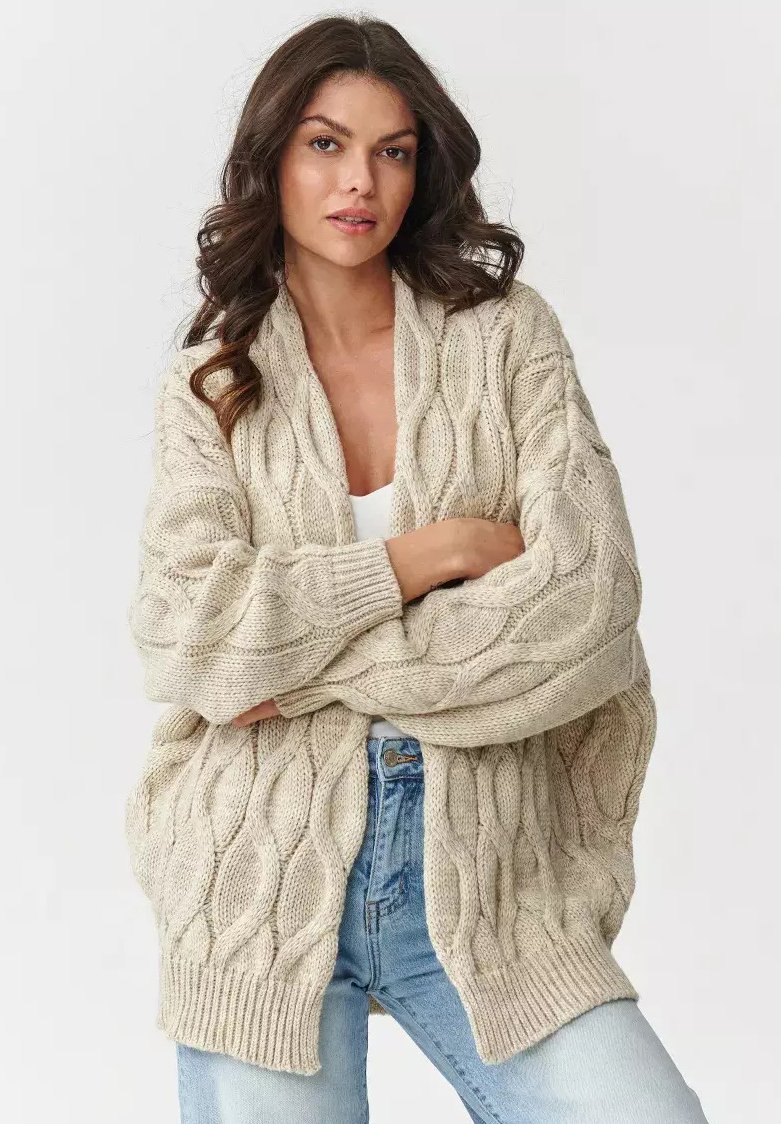 Kremowy kardigan z warkoczowym splotem, z kołnierzem szalowym i luźnym krojem, z prążkowanymi mankietami i dołem. Noszony na białej bluzce i jasnoniebieskich dżinsach.
