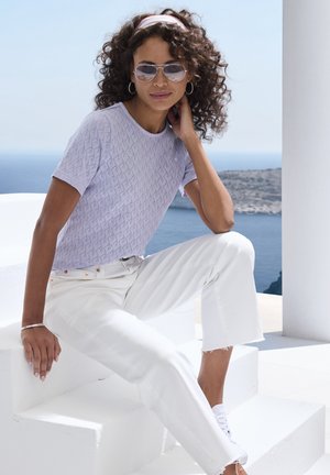 Haut tricot lilas à manches courtes avec un motif en losange, associé à un jean blanc court et des baskets blanches, sur fond côtier.