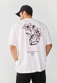 White cotton t-shirt with a loose fit, featuring black and pink floral illustrations on the back and text detailing.
