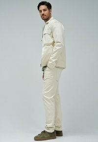 Beige Baumwolljacke mit Fronttaschen, kombiniert mit passender Hose. Das Model trägt dunkelgrüne Wildledersneaker und steht vor einem hellen Hintergrund.