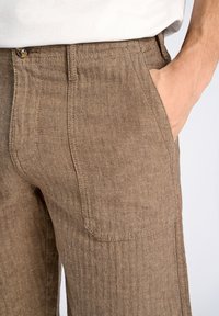 Pantalones de espiga marrones con una tela texturizada, bolsillos frontales y cierre de botón. El corte es entallado con un ajuste recto.