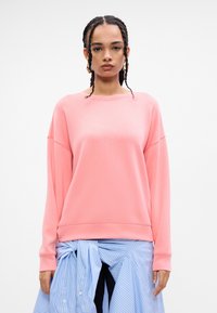 Koralrød genser med lange ermer, rund hals og ribbestrikket hem; kombinert med et blått og hvitt stripete skjørt som er knyttet i livet.