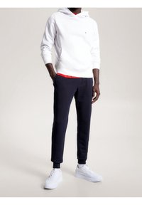 Felpa bianca con cappuccio, foderata di rosso, e logo; pantaloni della tuta blu navy; sneakers bianche con suole testurizzate; vestibilità rilassata.
