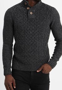 Pull en tricot gris foncé avec un motif texturé, un col haut avec deux boutons ronds en bois, et des poignets et un ourlet côtelés.