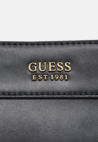 Surface en cuir noir avec des lettres métalliques dorées épelant "GUESS EST 1981" cousues dans le matériau.