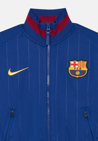 Veste zippée bleu avec des rayures verticales, col côtelé rouge, logo Nike doré et écusson brodé du FC Barcelone sur la poitrine gauche.