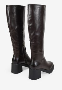 Botas de cuero marrón hasta la rodilla con una textura suave, tacón cuadrado y detalles de cremallera lateral. Punta redondeada y acabado pulido.