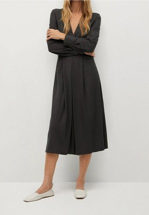 Frau trägt ein langärmeliges, schwarzes Midikleid und weiße flache Schuhe, steht mit verschränkten Armen vor einem schlichten hellen Hintergrund.