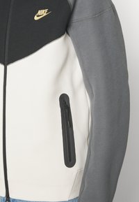 Giacca Nike con un design a blocchi di colore in nero, grigio e bianco, dotata di una tasca con zip e un grande logo dorato sul petto.