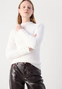 Weißer, flauschiger Strickpullover mit hohem Kragen und goldenen Knopfdetails an den Bündchen, kombiniert mit glänzenden, dunkelburgunderfarbenen eng anliegenden Hosen.