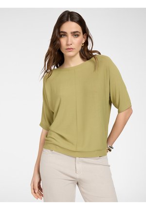 Donna con capelli castani lunghi che indossa una maglietta verde oliva a maniche corte e pantaloni beige chiaro, su sfondo bianco neutro.