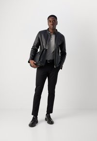 Veste en cuir noire sur un t-shirt henley gris, associée à un pantalon noir et des chaussures à lacets. Le sujet tient un sac noir, se tenant sur un fond clair.