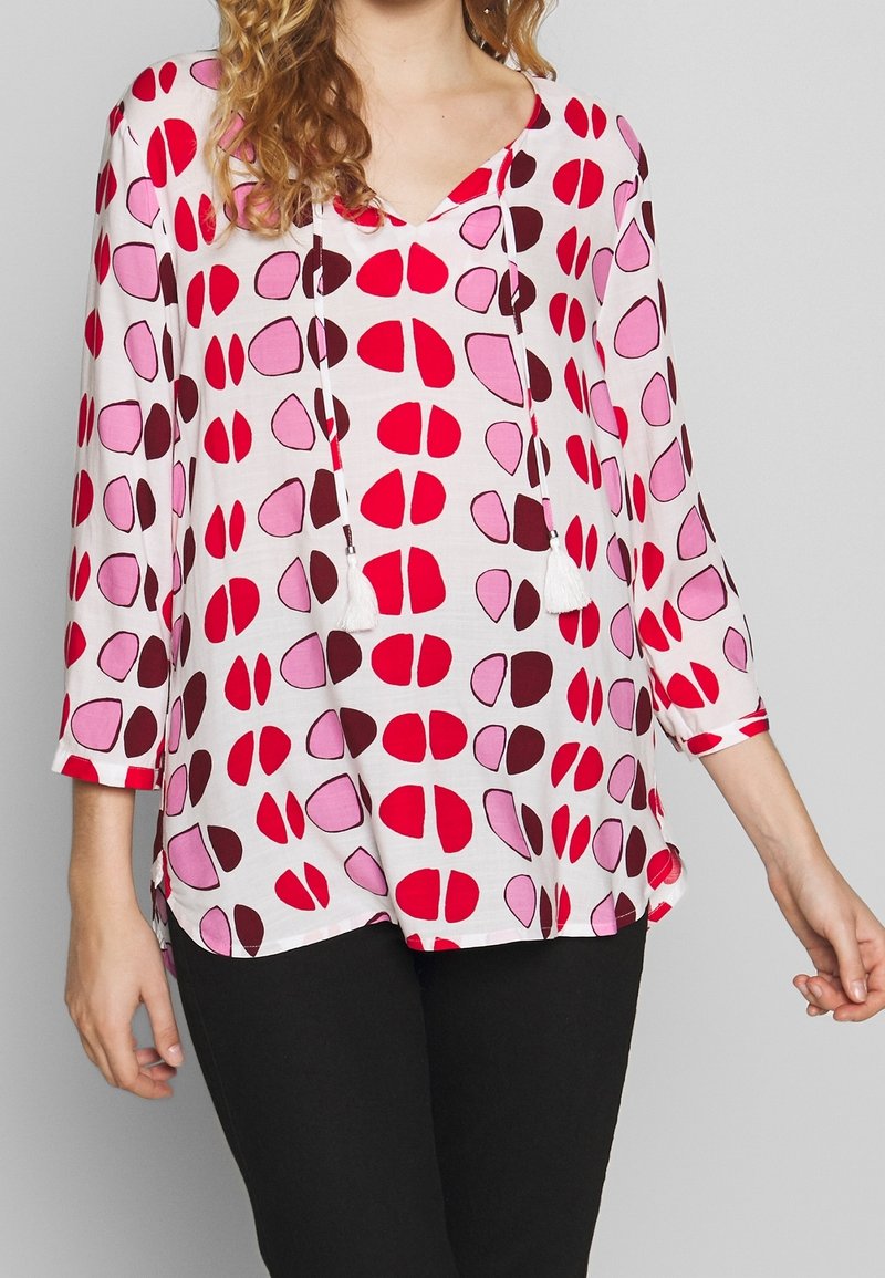 Haut léger, rose et blanc avec un décolleté en V, des manches 3/4 et des formes abstraites rouges et roses foncé. Doté d'un cordon de serrage avec des pompons.