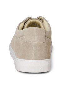 Zapato de ante beige con una textura suave, suela de goma blanca y cuello acolchado. Presenta un diseño minimalista con una lengüeta trasera para facilitar el uso.
