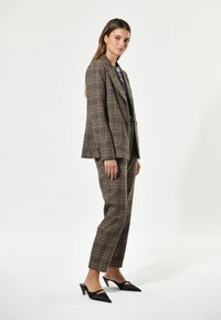 Abito a quadri marrone con blazer strutturato e pantaloni affusolati, caratterizzato da una consistenza liscia. Le mule nere con punta appuntita completano l'aspetto.
