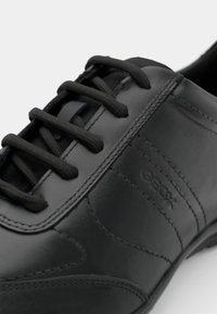 Chaussure en cuir noir avec une texture lisse, présentant un design basse, un bout rond et des lacets noirs. Logo embossé sur le côté.