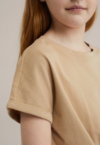 Béžové bavlněné tričko s kulatým výstřihem a ohrnutými krátkými rukávy. Měkká textura a jednoduchý design, přiléhavý, ale uvolněný střih.