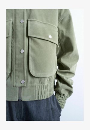 Veste en daim vert clair avec grandes poches à rabat sur le devant, boutons-pression argentés, poignets élastiques, et pantalon gris partiellement visible.