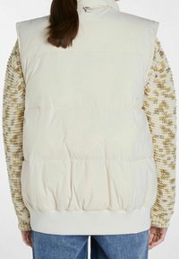 Polstrovaná krémová vesta s vysokým límcem, ribbovaným lemem a zapínáním na knoflíky, vrstvená přes strukturovaný svetr v zemitých tónech.