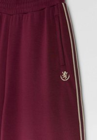 Bordowe spodnie z elastycznym pasem, be żowymi paskami po bokach, zamykana kieszonka oraz beżowy herb lwa w pobliżu kieszeni po prawej stronie.