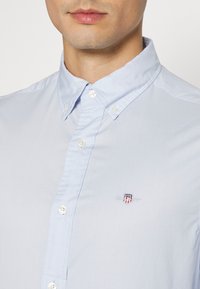 Homme portant une chemise bleu clair à boutons avec un petit logo écusson brodé en rouge, blanc et bleu sur la poitrine.