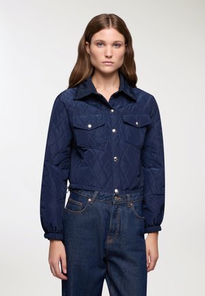 Giacca trapuntata blu navy con chiusura a bottoni, colletto a punta e due tasche sul petto. I polsini elastici e la vestibilità corta si abbinano ai jeans in denim scuro.