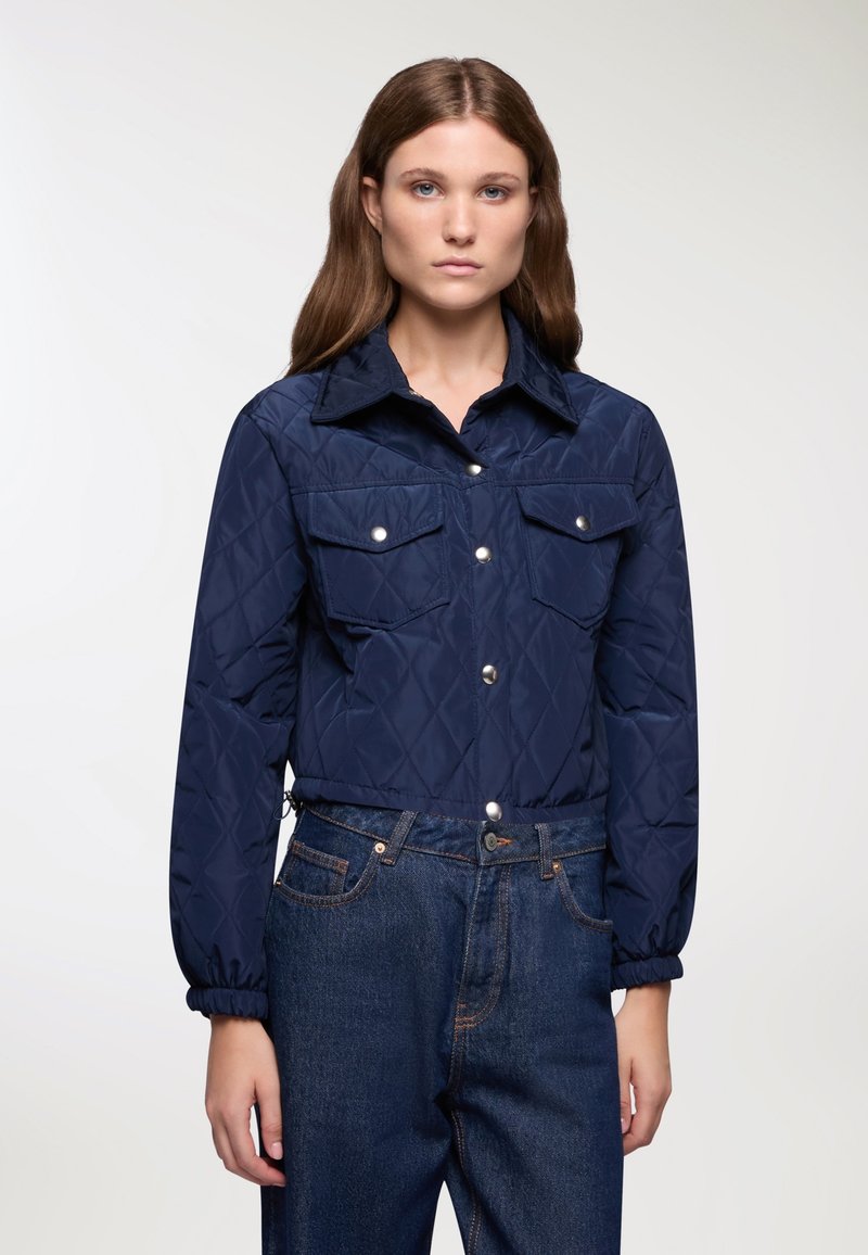 Giacca trapuntata blu navy con chiusura a bottoni, colletto a punta e due tasche sul petto. I polsini elastici e la vestibilità corta si abbinano ai jeans in denim scuro.