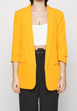Persona que lleva una chaqueta blazer amarilla brillante sobre un top corto blanco, pantalones negros de talle alto con un cinturón de hebilla grande, y tatuajes visibles en la mano y el pecho.