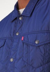 Mörkblå vadderad nylonjacka med tryckknappskrage, två bröstfickor med klaffar och en liten röd Levi's-logotag.