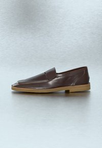 Mocassin à enfiler en cuir brun foncé avec une semelle en caoutchouc beige, un talon bas et des coutures minimales sur un fond gris clair.