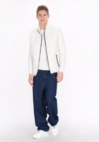 Weiß gesteppte Weste mit Reißverschluss und navyfarbenen Akzenten, getragen über einem weißen Langarmshirt und kombiniert mit locker sitzenden blauen Jeans und weißen Sneakern.