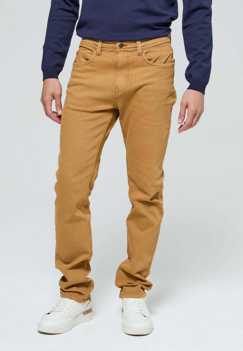 Jean en denim beige avec une coupe droite, dotée de poches avant et d'une fermeture à bouton en métal. Associé à des baskets blanches et un pull bleu marine.