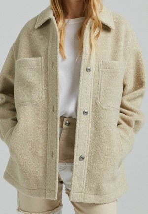 Vrouw draagt een beige wollen jas met knopen en grote borstzakken over een wit shirt en lichtbeige broek met rafelige zomen.