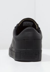 Black leather sneaker with a flat sole, rounded back, and prominent Adidas logo on the heel. Interior shows a soft lining and label.