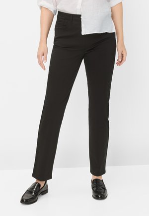 Persoon die strakke zwarte broek, zwarte loafers en een wit overhemd met opgerolde mouwen draagt, staand tegen een effen witte achtergrond.
