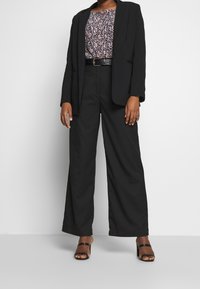 Blazer noir ajusté sur un haut à motifs, associé à un pantalon noir taille haute et à pattes d'éléphant, et des talons aiguilles noirs à lanières.