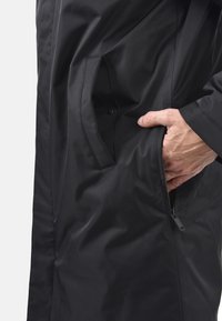 Giacca nera impermeabile con texture liscia, dettagli delle tasche laterali e chiusura con zip, che mostra un design moderno e minimale.