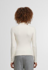 Langärmliges, hochgeschlossenes Oberteil in Off-White, aus weichem, dehnbarem Material. Es bietet eine taillierte Passform und eine glatte Textur, kombiniert mit grauen Hosen.