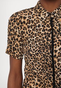 Skjorte med leopardmønster og korte ermer, med en svart knappestolpe foran og kontrastfarge på kragen. Teksturert stoff i brune og svarte toner.