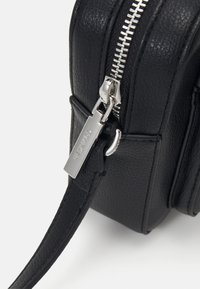 Schwarze Ledertasche mit strukturierter Oberfläche, ausgestattet mit einem silbernen Reißverschluss, einem Hardware-Tag und einem dünnen Schultergurt. Kompaktes und rechteckiges Design.