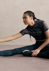 Haut de sport noir à motifs et leggings, fabriqués dans un matériau extensible, au design ajusté. Une personne s'étire sur un sol clair.