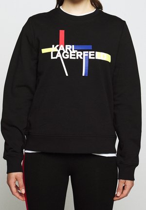 Czarna bluza z nadrukiem „KARL LAGERFELD” na czerwonych, niebieskich, żółtych i białych abstrakcyjnych liniach noszona przez osobę.