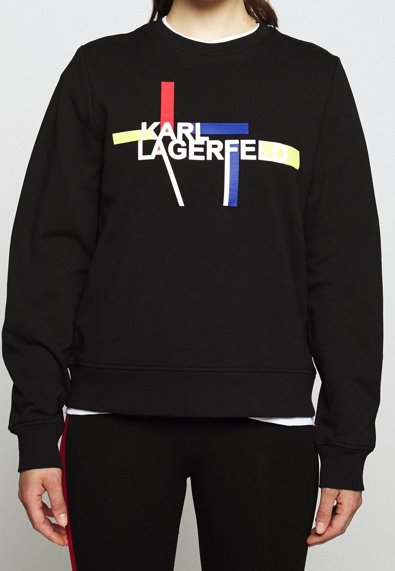 Sweat-shirt noir avec le texte « KARL LAGERFELD » superposé sur des lignes abstraites rouges, bleues, jaunes et blanches porté par une personne.