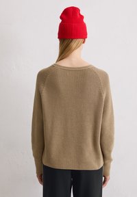 Beige gebreide trui met ribbelstructuur, brede halslijn en verlaagde schouders, gecombineerd met een rode ribbelgehaakte muts. Zwarte broek eronder.