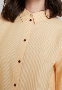 Gestreepte gele shirt met een pointed collar, bruine knopen en een gladde textuur. Bevat verticale witte strepen voor extra detail.