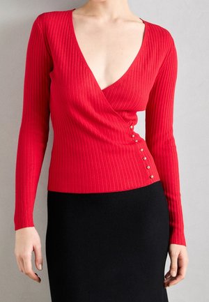 Femme portant un pull cache-cœur rouge à manches longues côtelé avec des boutons sur le côté, associé à une jupe noire.